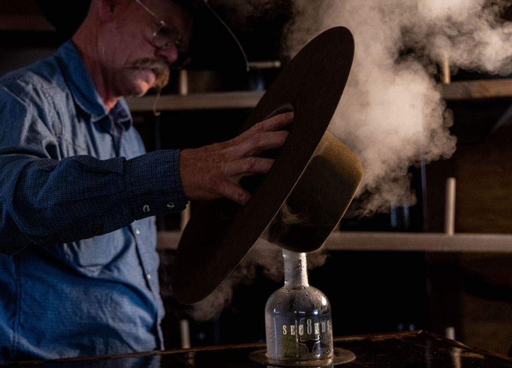 A man, Chaz Mitchell, standing in front of a hat steamer, steaming a brown cowboy hat.